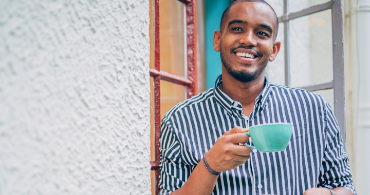 Man standing outside his doorway, smiling peacefully while enjoying a hot morning beverage in bright, natural light, capturing a calm and joyful start to the day.